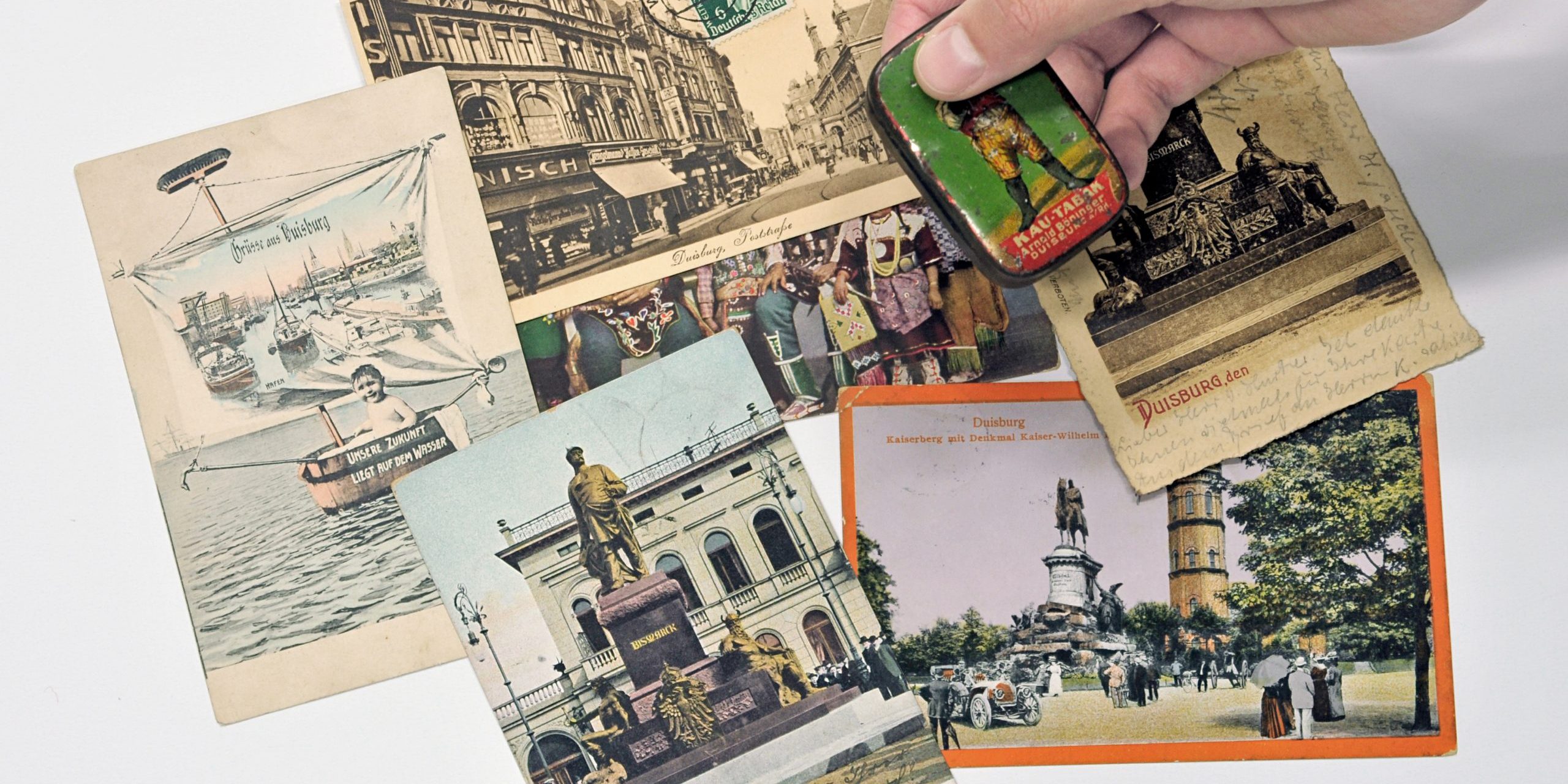 Verschiedene Postkarten mit Abbildungen des Bismarck-Denkmals in Duisburg. Außerdem eine historische Tabakdose der Firma Böninger mit rassistischem Werbemotiv.