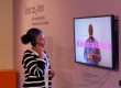 Eine junge Schwarze Frau hört sich ein Erklärvideo zum Thema "Kolonialismus" in der Ausstellung Überseehen an.