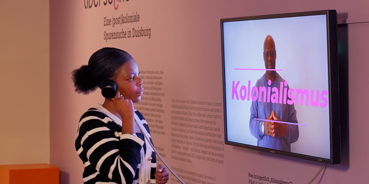 Eine junge Schwarze Frau hört sich ein Erklärvideo zum Thema "Kolonialismus" in der Ausstellung Überseehen an.