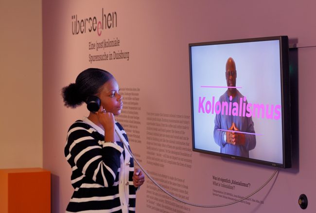 Eine junge Schwarze Frau hört sich ein Erklärvideo zum Thema "Kolonialismus" in der Ausstellung Überseehen an.