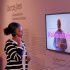 Eine junge Schwarze Frau hört sich ein Erklärvideo zum Thema "Kolonialismus" in der Ausstellung Überseehen an.
