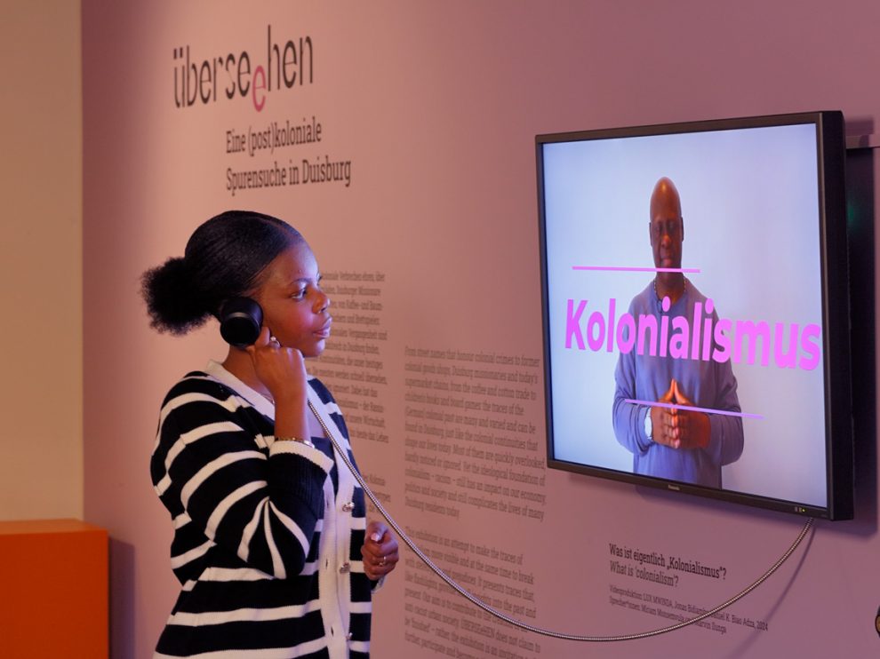 Eine junge Schwarze Frau hört sich ein Erklärvideo zum Thema "Kolonialismus" in der Ausstellung Überseehen an.