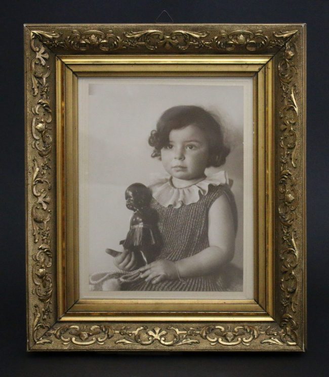 Kinderfoto von Inge Stern, ca. zwei-jähriges Mädchen sitzt mit ernstem Gesicht aufrecht, mit Schwarzer Puppe in der Hand; Schwarz-weiß-Fotografie in goldenem Rahmen; Inventarnummer 2020:0019; Sammlung ZfE, Kultur- und Stadthistorisches Museum Duisburg