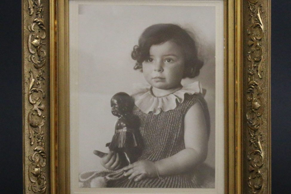 Kinderfoto von Inge Stern, ca. zwei-jähriges Mädchen sitzt mit ernstem Gesicht aufrecht, mit Schwarzer Puppe in der Hand; Schwarz-weiß-Fotografie in goldenem Rahmen; Inventarnummer 2020:0019; Sammlung ZfE, Kultur- und Stadthistorisches Museum Duisburg