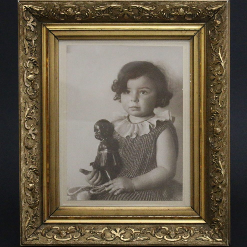 Kinderfoto von Inge Stern, ca. zwei-jähriges Mädchen sitzt mit ernstem Gesicht aufrecht, mit Schwarzer Puppe in der Hand; Schwarz-weiß-Fotografie in goldenem Rahmen; Inventarnummer 2020:0019; Sammlung ZfE, Kultur- und Stadthistorisches Museum Duisburg