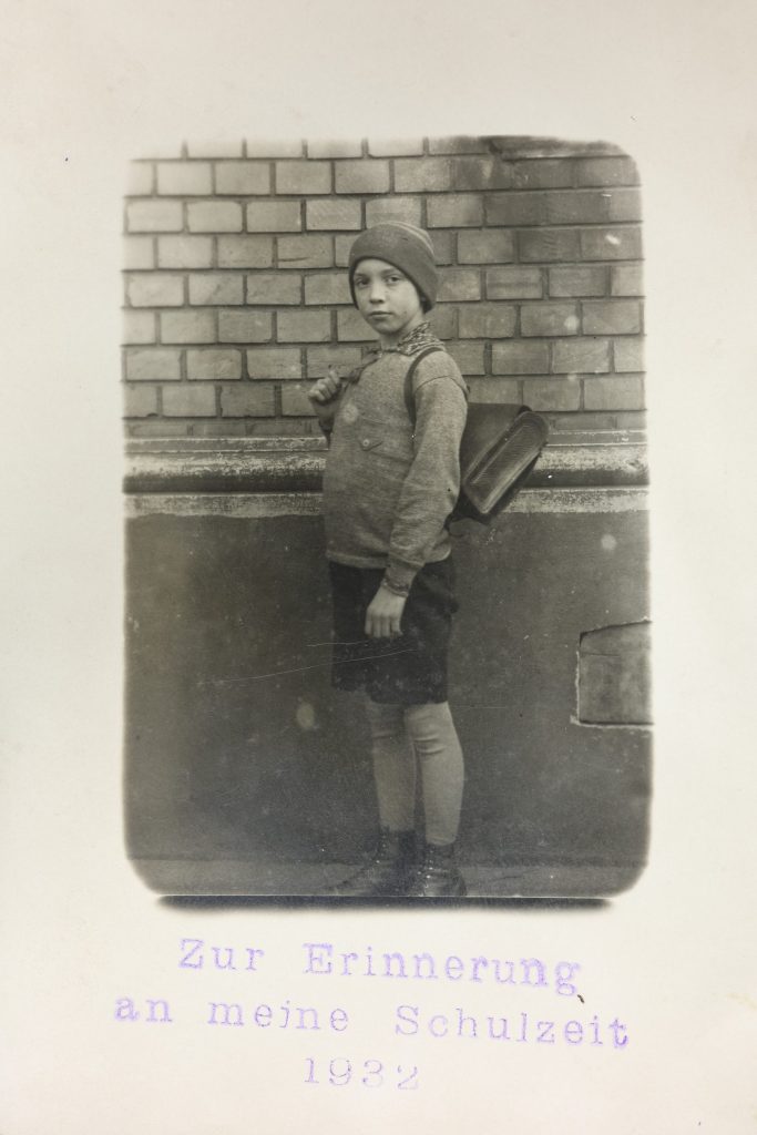 Abb. 1 alternati_ Das Foto zeigt den 10-jährigen Karl Heinz im 4. Volksschuljahr der Carpschule und das Gebäude des Realgymnasiums, in dem er zwei Jahre später die Zeichnungen anfertigen musste. Er trägt einen ledernen Schulranzen, kurze Hosen, darunter eine Strumpfhose und hoch geschnürte Lederschuhe. Auf dem Kopf ist eine Mütze.
